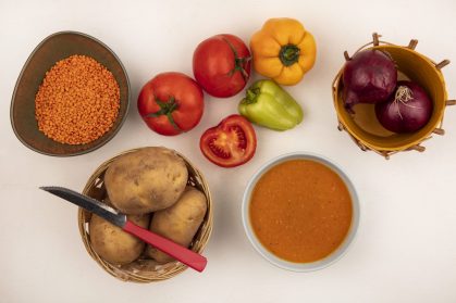 top view of nutritious lentil soup on a bowl with red onions on a bucket with potatoes on a bucket with knife with peppers and tomatoes isolated on a white background