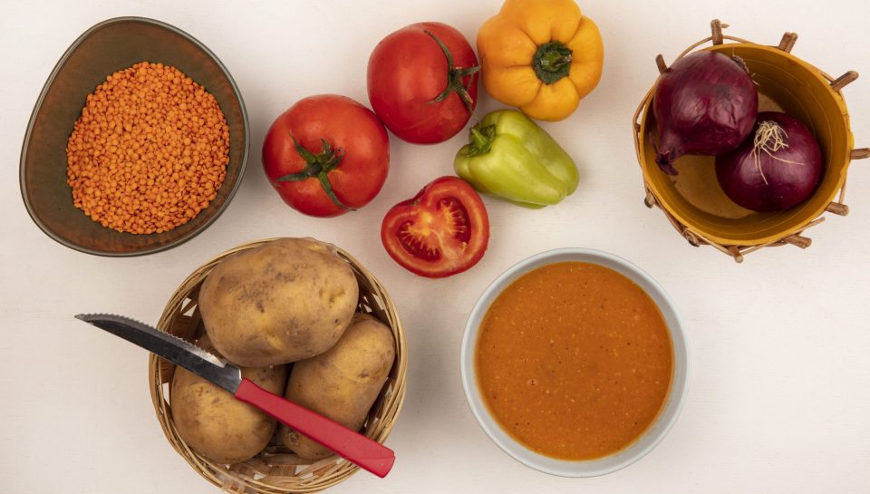 top view of nutritious lentil soup on a bowl with red onions on a bucket with potatoes on a bucket with knife with peppers and tomatoes isolated on a white background